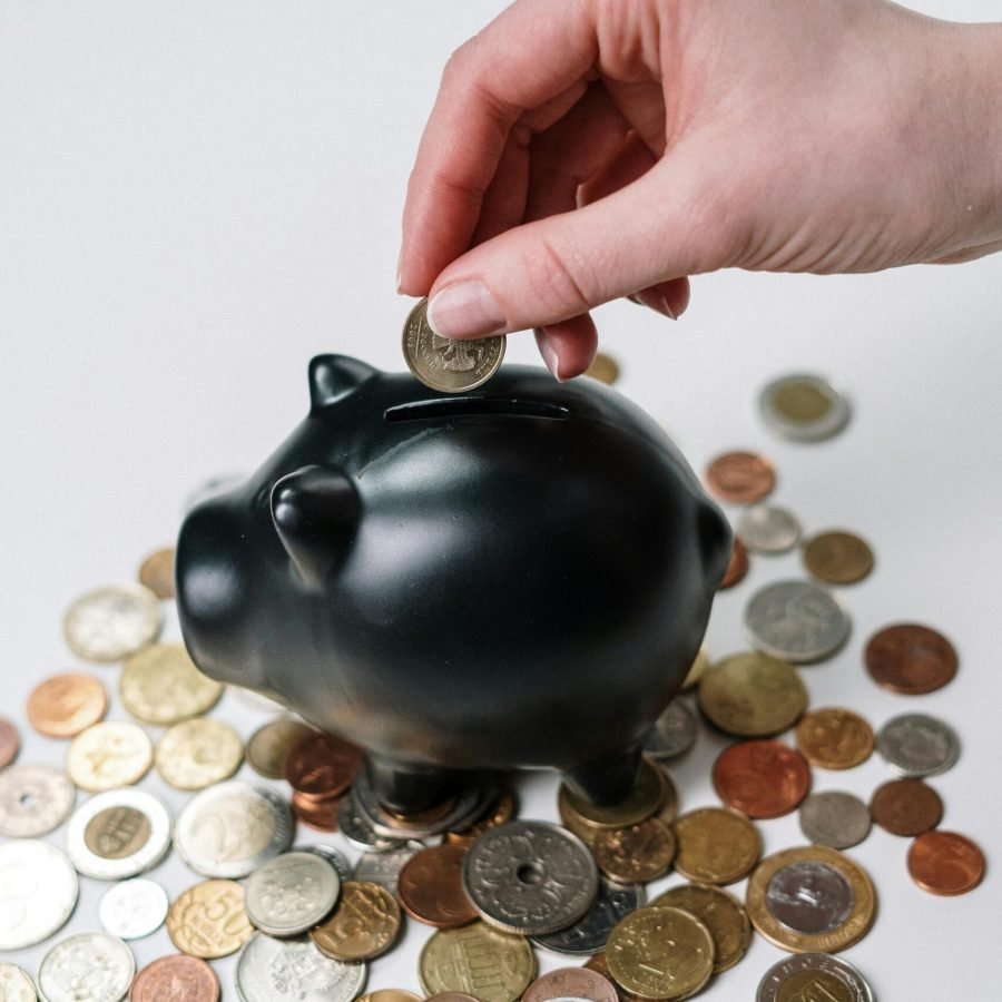 pexels-photo-3943723-3943723 Hand inserting coin into a black piggy bank surrounded by various coins, symbolizing savings.